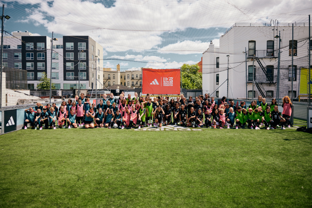 More than 100 girls pose for a group photo in front of a goal and sign with the adidas and U.S. Soccer Foundation logos on a soccer pitch